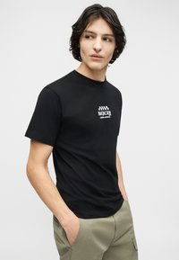 Νεαρός με σκοτεινά μαλλιά που φοράει μαύρο μπλουζάκι με στάμπα και ανοιχτόχρωμο ελιάς παντελόνι cargo, κοιτάζοντας προς τα πλάγια με τα χέρια στις τσέπες.