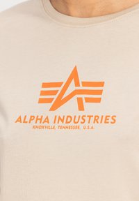 Beige bavlněné tričko s oranžovým grafickým potiskem "ALPHA INDUSTRIES" a logem. Kulatý výstřih a hladká textura.