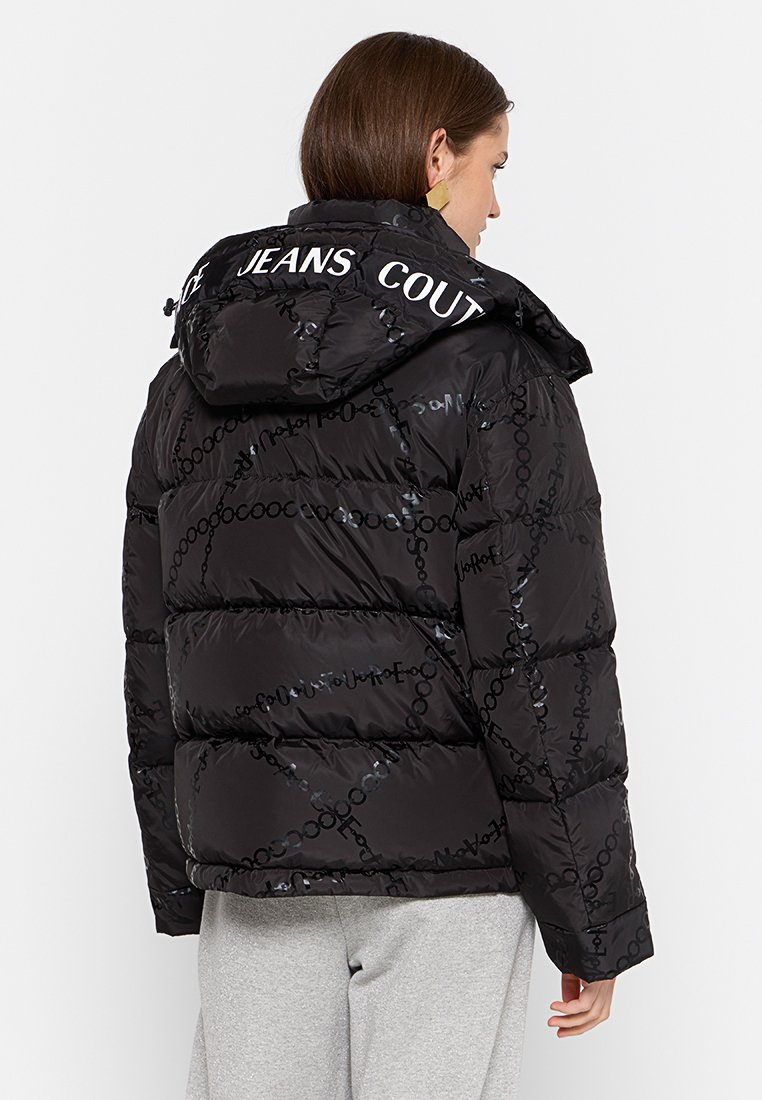 Mujer con una chaqueta acolchada negra con un patrón de cadenas y el texto "COUTURE JEANS" en la capucha, combinada con pantalones gris claro, de espaldas.