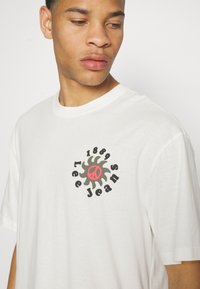 Camiseta de algodón blanca con un gráfico circular que dice "Lee Jeans 1889" en negro y verde, con un símbolo de paz rojo en el centro. Mangas cortas.