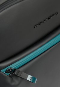 Superficie de cuero negro con una cremallera turquesa, una lengüeta de cuero para tirar y el nombre de la marca "piquadro" en relieve.