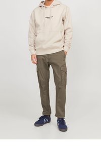 Beige huvtröja med framficka och logotyp, tillsammans med olivgröna cargobyxor med sidofickor och mörkblå sneakers.
