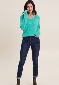 Türkiser gestrickter Pullover mit V-Ausschnitt, ausgestattet mit einem strukturierten Muster. Kombiniert mit dunklen Skinny Jeans und Stiefeletten.