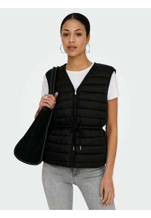 Donna che indossa un gilet trapuntato nero senza maniche sopra una maglia bianca a maniche corte, jeans grigi, tiene una borsa nera a tracolla e indossa orecchini a cerchio.