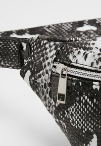 Banane à motif peau de serpent noir et blanc avec une fermeture éclair argentée et une languette rectangulaire, sur un fond clair uni.