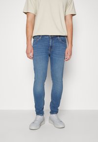 Vaqueros de mezclilla azul de corte ajustado con cinco bolsillos, sutiles desgastes en los muslos y cremallera. Combinados con zapatillas deportivas blancas.