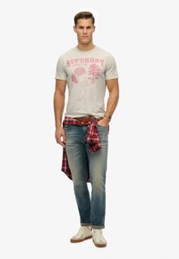 Camiseta gris con estampado gráfico rojo, jeans azules y una camisa de cuadros roja atada a la cintura. Zapatillas beige completan el atuendo.