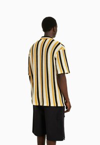 Koszula z krótkim rękawem w żółte, czarne i białe pionowe paski. Wykonana z teksturowanej tkaniny, z kołnierzykiem i luźnym krojem.
