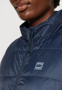 Granatowa kurtka puchowa z wysokim kołnierzem, zamek błyskawiczny i naszywka z logo GAP. Wykonana z gładkiego, błyszczącego materiału o pikowanej strukturze.