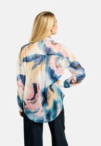 Szőke nő, kezét csípőjén tartva, laza, hosszú ujjú blúzt visel, amely absztrakt rózsaszín, kék, barack és fehér csavarmintával, valamint fekete nadrággal.