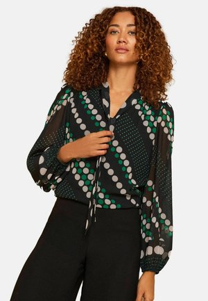 Nainen kiharatukkainen pukeutuneena mustaan paitaan, jossa on vihreitä ja valkoisia pilkkuja, sekä mustiin leveälahkeisiin housuihin. Hän poseeraa käsi puseron kauluksen lähellä.