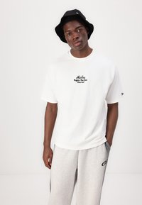 White cotton t-shirt with black embroidered text, paired with light grey sweatpants featuring a dark logo, and a black bucket hat.