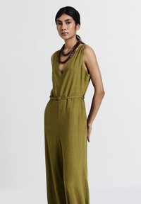 Jumpsuit senza maniche color verde oliva con scollatura a V, vita con cintura e gamba larga. Modella accessoriata con una collana di perle marroni a strati.