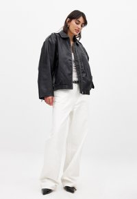 Giacca in pelle nera cropped con colletto alto, chiusura con zip e polsini elasticizzati, abbinata a pantaloni bianchi a gamba larga e tacchi neri appuntiti.