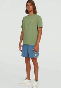 Man with shoulder-length hair wearing green short-sleeve shirt, blue patterned shorts, and white sneakers standing on plain background.