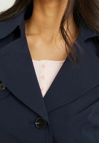 Granatowy płaszcz trench z dużym kołnierzem, detalami guzika oraz kontrastującą jasnoróżową bluzką z prążkowanego materiału z guzikami. Gładka tekstura, dopasowany krój.