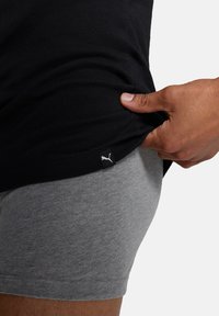 Schwarzes Baumwoll-T-Shirt mit einem grauen Taillenband, das ein kleines weißes Puma-Logo-Tag zeigt. Der Stoff wirkt weich und enganliegend.