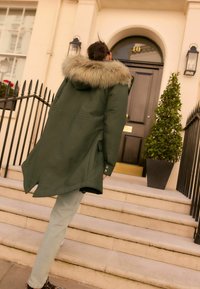 Grüne Parka mit pelzgefütterter Kapuze, Merkmale einer taillierten Taille, Taschen und einem längeren hinteren Saum, kombiniert mit hellblauen Jeans und gemusterten Schuhen.