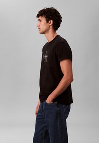 Crna pamučna majica kratkih rukava, s okruglim izrezom i bijelim Calvin Klein logotipom na prsima, u kombinaciji s plavim trapericama.