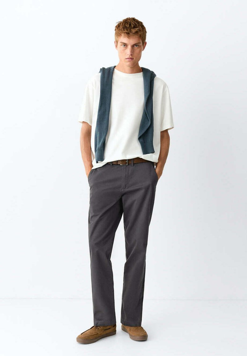 Jeune homme debout, les mains dans les poches, portant un t-shirt blanc, un pantalon foncé, des chaussures marron et un pull sombre drapé sur les épaules.