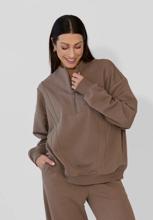 Kobieta ubrana w brązową bluzę zapinaną na zamek oraz pasujące spodnie, uśmiechająca się podczas regulowania zamka jedną ręką, a druga trzymająca się za kieszeń.