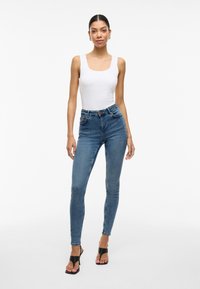 Bodysuit branco sem mangas combinado com jeans skinny azuis de cintura alta. Modelo a usar sandálias de salto abertas pretas. Tecido suave, design ajustado.