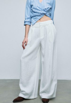 Kvinde iført løstsiddende, hvide vidde bukser, blåstribet cropped skjorte og brune spidse sko, stående på grå gulv mod en ensfarvet væg.