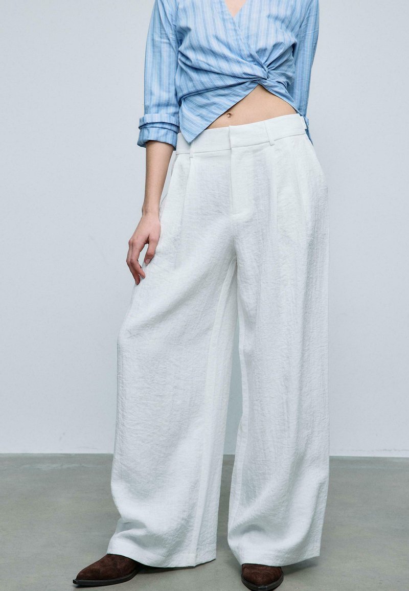 Kvinde iført løstsiddende, hvide vidde bukser, blåstribet cropped skjorte og brune spidse sko, stående på grå gulv mod en ensfarvet væg.