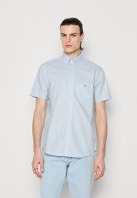 Homme aux cheveux plaqués en arrière, portant une chemise bleue claire à manches courtes boutonnée et un jean bleu clair, debout devant un fond blanc uni.