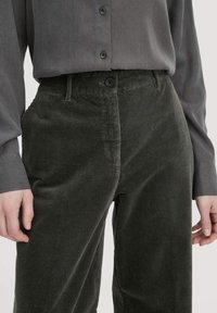 Hoge taille donkergroene corduroy broek met knoop- en ritssluiting, gecombineerd met een ingestopte lange mouwen grijze blouse.