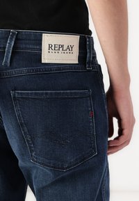 Tamno plave traperice s vidljivom kožnom etiketom na pojasu koja glasi "REPLAY" i detaljnim šavom na stražnjem džepu.