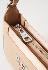 Μπεζ ψεύτικη δερμάτινη τσάντα με ασημένιες λεπτομέρειες και κλείσιμο φερμουάρ. Ο εσωτερικός χώρος έχει υφή επένδυσης και ανάγλυφη λεπτομέρεια λογότυπου.