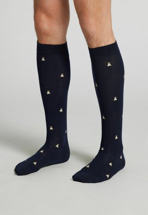 Nohy obuté v tmavě modrých ponožkách sahajících nad kolena s malými vzory medvídků stojící na šedém povrchu.