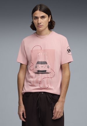 Unge mand med skulderlangt mørkt hår iført en lyserød T-shirt med en hvid Porsche-bil grafik og "9" på ærmet.