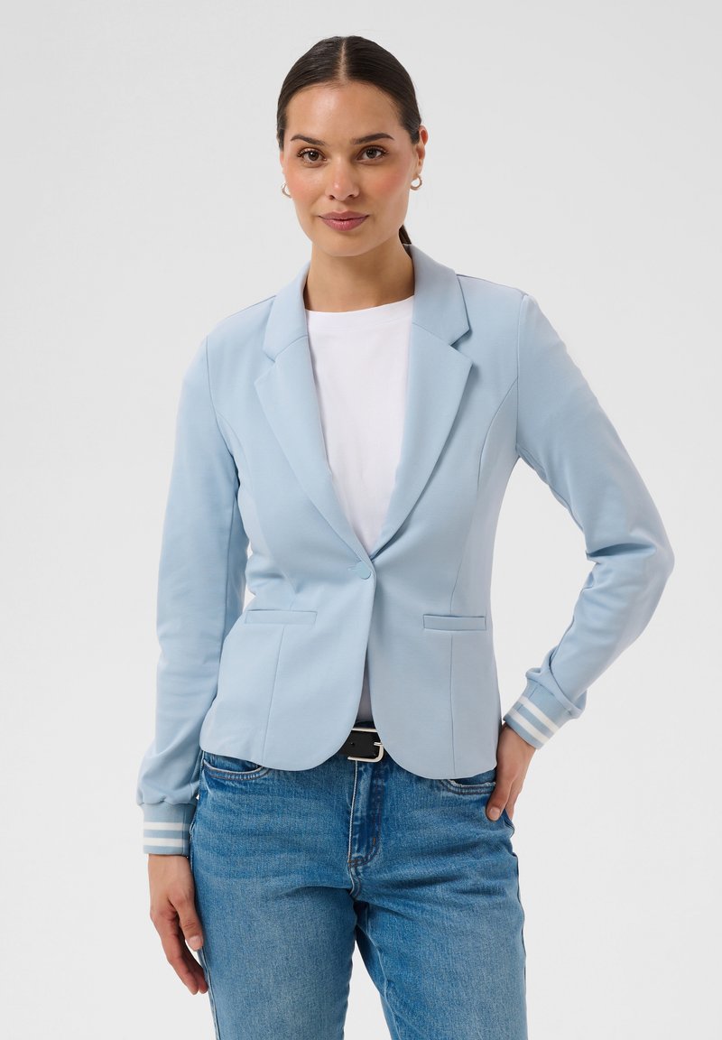 Femme debout, portant un blazer bleu clair sur une chemise blanche et un jean bleu, une main dans la poche, fond neutre.