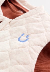 Gros plan sur un tissu matelassé beige avec un détail de visage souriant brodé en bleu, reposant sur un tissu côtelé couleur rouille.