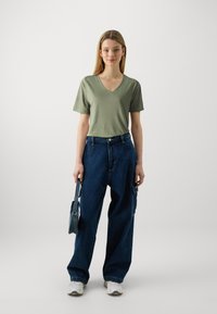 T-shirt vert olive à manches courtes et col en V associé à un pantalon cargo en denim bleu ample. Le mannequin tient un petit sac à main bleu. Des baskets blanches complètent le look.
