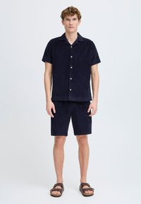 Marineblå kordfløyelskortermet skjorte og matchende shorts, med krage, frontknapper og en teksturert finish. Brune sandaler fullfører antrekket.