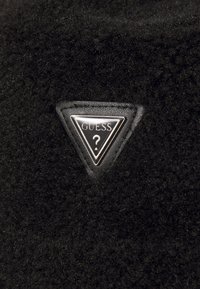 Tessuto di pile nero con una toppa triangolare in pelle nera che presenta un logo "GUESS" in argento e un punto interrogativo al centro.