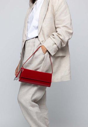 Mujer que lleva un traje beige claro y una camiseta blanca sosteniendo un bolso de mano rojo brillante rectangular con una correa delgada.
