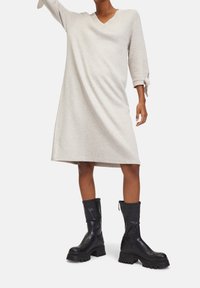 Personne portant une robe midi beige clair avec des manches trois-quarts et des poignets noués, associée à des bottes noires épaisses mi-mollet.