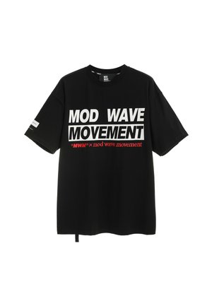 Joven con camiseta negra "Mod Wave Movement", shorts vaqueros azul claro con parche de oso, calcetines blancos y zapatos negros texturizados, de pie.