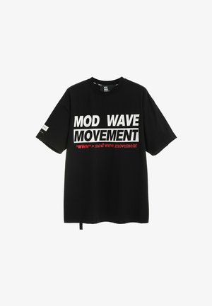 Joven con camiseta negra "Mod Wave Movement", shorts vaqueros azul claro con parche de oso, calcetines blancos y zapatos negros texturizados, de pie.