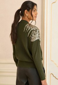 Olīvzaļš džemperis ar brīvu piegriezumu, ar metalizētām pērlītēm uz vienas pleca un ribotām piedurknēm. Gluda tekstūra.