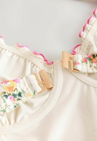 Beige stoffen top met roze rimpelige randen. Bloemenpatroon op de bandjes met gouden hardware voor het bevestigen. Gladde textuur, ronde halslijn.