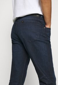 Mörkblå denimjeans med slim fit, med bakfickor, diskret sömnad och en liten märkesetikett nära midjan.