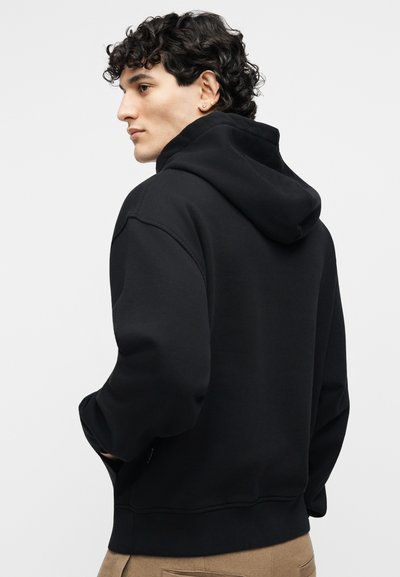 Jaunas žmogus su garbanotais tamsiais plaukais, dėvintis juodą gobtuvą ir smėlio spalvos kelnes, šiek tiek pasisukęs į šoną priešais paprastą foną.