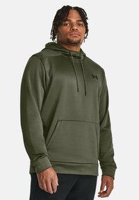 Olivengrøn hoodie lavet af glat materiale, med snore i hætten, frontlomme og subtil sort logo på brystet.
