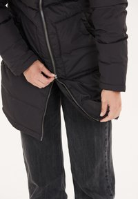 Sort dunjakke med quiltet tekstur, lynlås foran og sidelommer; model justerer lynlåsen mens han har mørke jeans på.