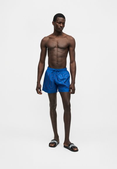 Λεπτός άνδρας που στέκεται, φοράει μπλε μπλε μαγιό με το κείμενο "MOSCHINO" και μαύρα σανδάλια slip-on με λευκή επιγραφή "MOSCHINO" σε μονόχρωμο φόντο.